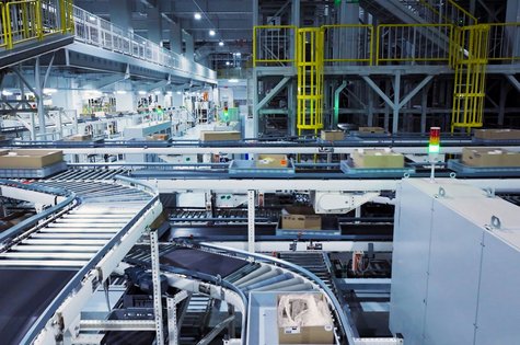 In einer großen Lagerhalle sind Förderbänder zu sehen, die Kartons transportieren. Mehrere Ebenen und Ausstattungsteile deuten auf ein automatisiertes Logistiksystem hin. Beleuchtung und Sicherheitsvorrichtungen sind vorhanden.