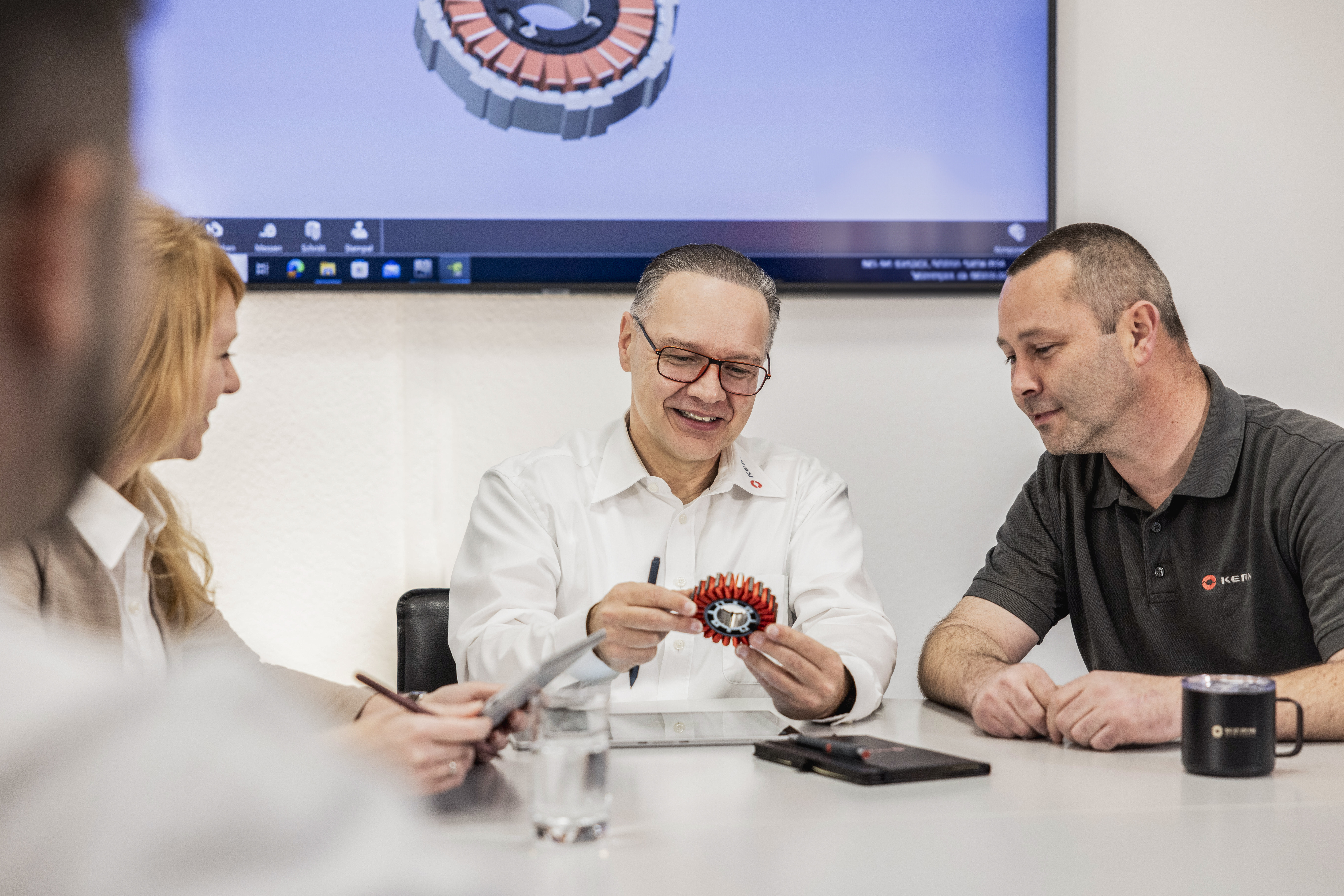 Vier Personen in einem Besprechungsraum, ein Mann in weißem Hemd präsentiert ein technisches Bauteil, im Hintergrund ein Bildschirm mit Firmenlogo.